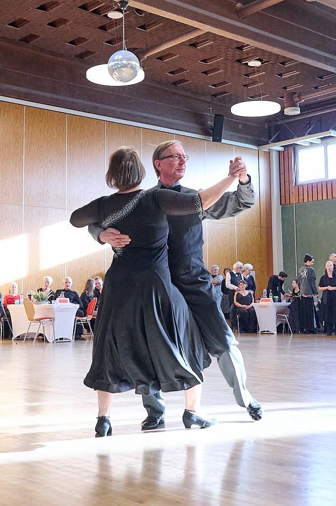 Tanzendes Paar in eleganter Kleidung auf einem Tanzboden, umgeben von anderen G&auml;sten in einem Saal.