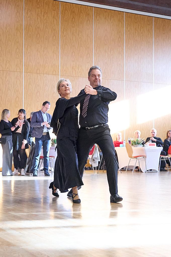 Tanzendes Paar in eleganter Kleidung auf einer Parkettfl&auml;che, umgeben von G&auml;sten bei einer Veranstaltung.