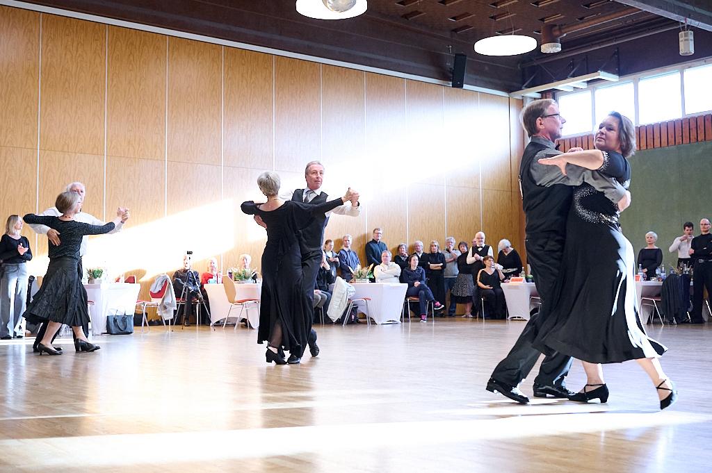 Paare tanzen in eleganter Kleidung auf einer Holzparkettfl&auml;che, w&auml;hrend Zuschauer in einem Festsaal zuschauen.