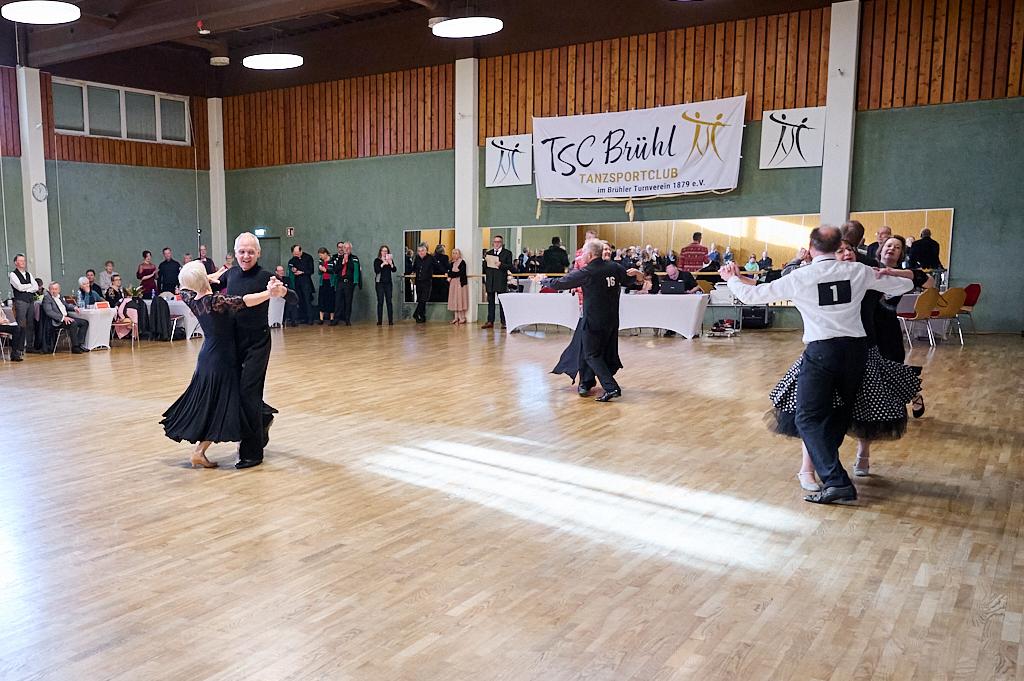 Tanzende Paare in eleganter Kleidung auf einer Holzfl&auml;che w&auml;hrend einer Veranstaltung im TSC Br&uuml;hl im Hintergrund.