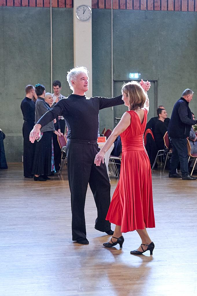 Tanzpaar in Aktion: Mann in Schwarz und Frau in rotem Kleid tanzen auf einem Parkettboden, im Hintergrund weitere T&auml;nzer.