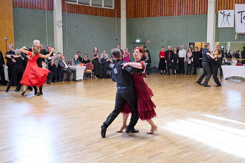 Tanzpaar in stilvoller Kleidung tanzt auf einer Holzfl&auml;che, umgeben von Zuschauern und weiteren T&auml;nzern.