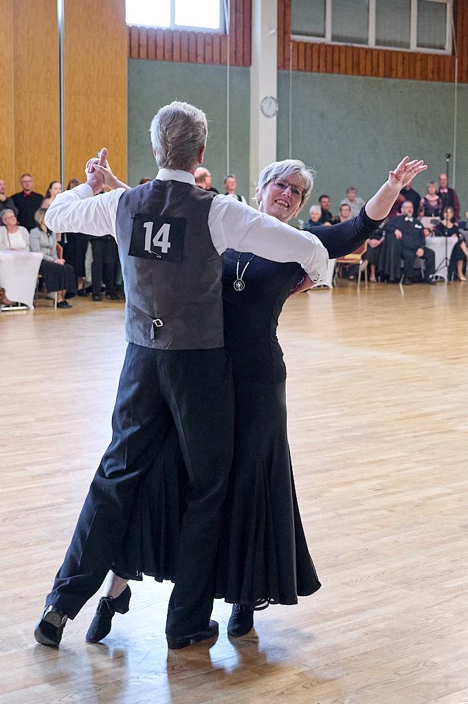Tanzpaar in eleganter Kleidung f&uuml;hrt einen Walzer auf einer Tanzfl&auml;che, Publikum im Hintergrund beobachtet.