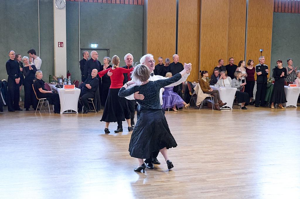 Tanzendes Paar in eleganter Kleidung auf einer Tanzfl&auml;che, umgeben von Zuschauern und Tischen im Hintergrund.
