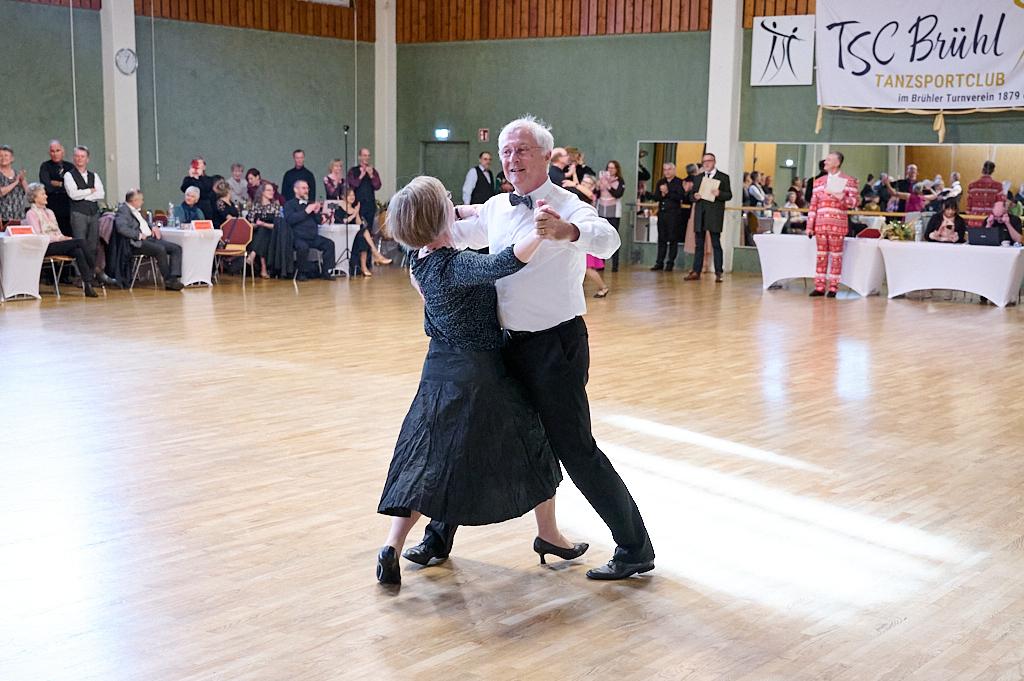 Tanzendes Paar in formeller Kleidung auf einer Tanzfl&auml;che, umgeben von Zuschauern und Tischen im Hintergrund.