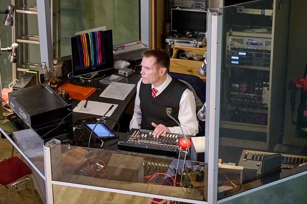 Mann in einem Kontrollraum sitzt vor Mischpult und Monitor, umgeben von technischen Ger&auml;ten und Kabeln.