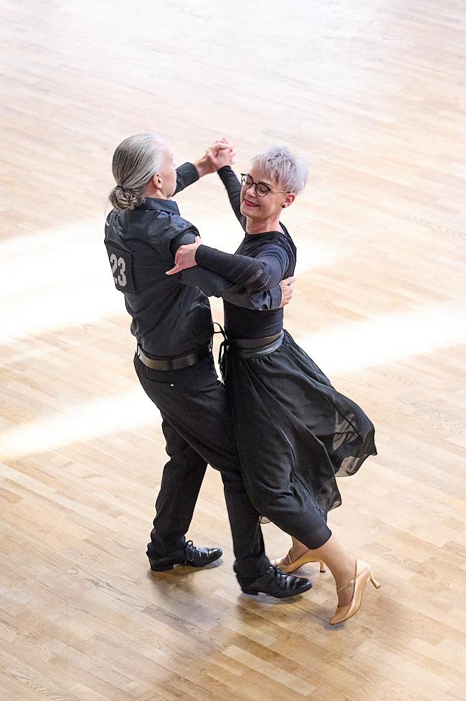 Tanzpaar in eleganter Kleidung, das sich auf einem hellen Parkettboden im Cha-Cha bewegt.