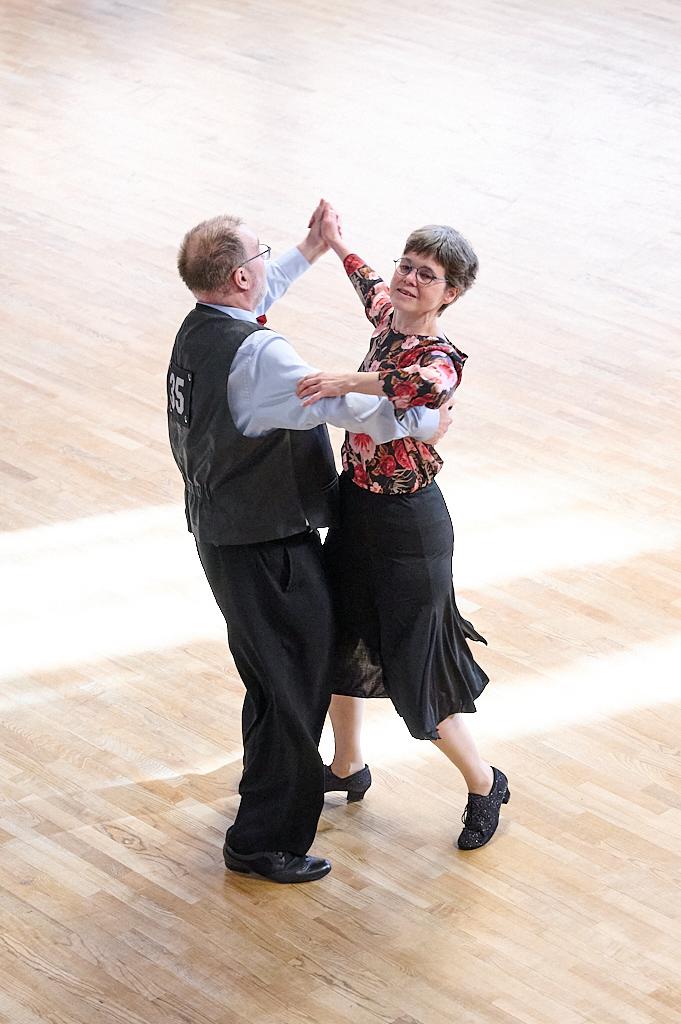 Tanzpaar in eleganter Kleidung, das eng umschlungen auf einem Holzboden tanzt. Helle Lichtstrahlen fallen ein.