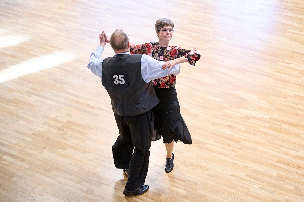 Tanzendes Paar in formeller Kleidung auf einem Holzboden, Mann mit Nummer 35 auf dem R&uuml;cken, Frau in bunter Bluse.