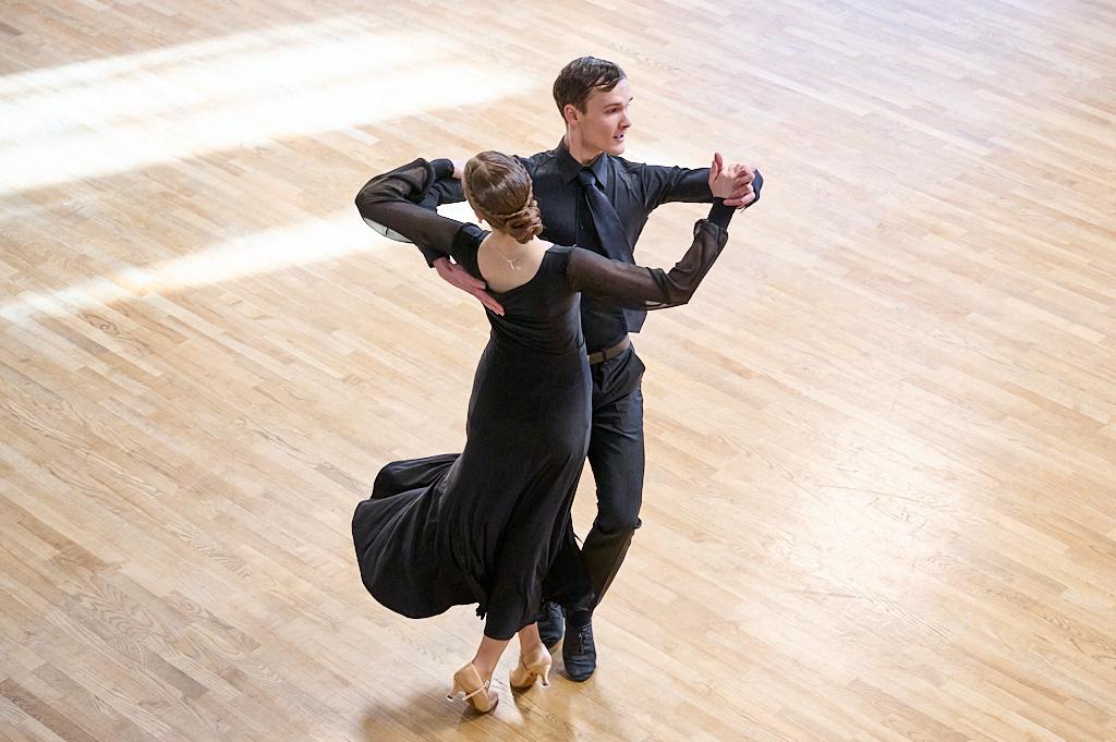 Tanzendes Paar in eleganter Kleidung auf einem hellen Holzboden, der m&auml;nnliche T&auml;nzer f&uuml;hrt die Dame in einem Schwarzkleid.