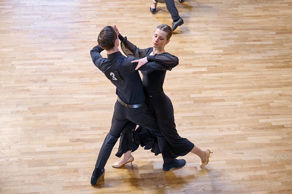 Tanzpaar in schwarzer Kleidung f&uuml;hrt einen Paartanz auf einem hellen Holzboden aus.