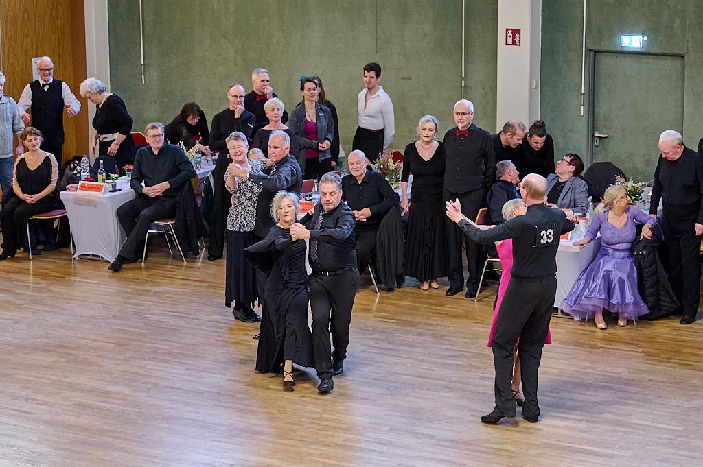 Tanzpaar in eleganter Kleidung auf einer Tanzveranstaltung, umgeben von Zuschauern an festlich gedeckten Tischen.
