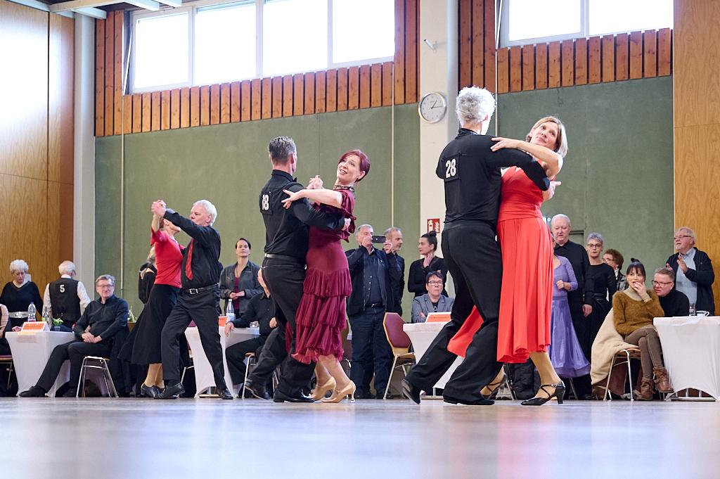 Tanzende Paare in eleganter Kleidung auf einer B&uuml;hne, Zuschauer sitzen an Tischen im Hintergrund.
