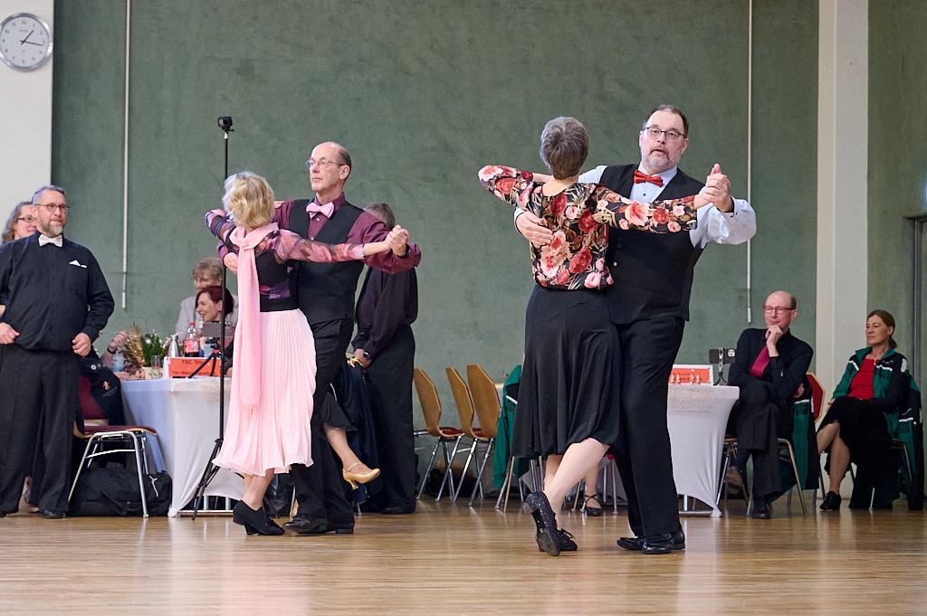 Tanzpaare in eleganter Kleidung bei einem Tanzwettbewerb auf einer Holzfl&auml;che, Publikum im Hintergrund.