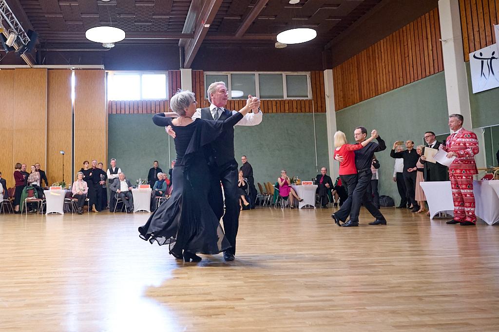 Tanzpaar wirbelt in eleganter Kleidung &uuml;ber eine Tanzfl&auml;che, Zuschauer applaudieren im Hintergrund.