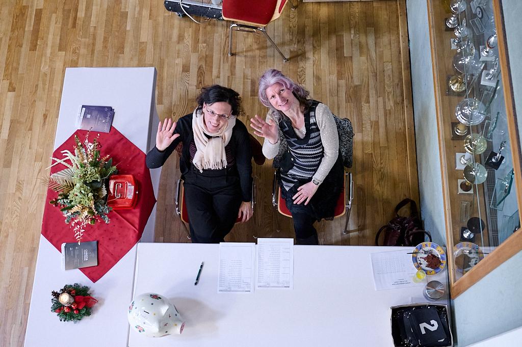 Zwei Frauen sitzen an einem Tisch, winken freundlich und umgeben von Dekoration und Unterlagen.