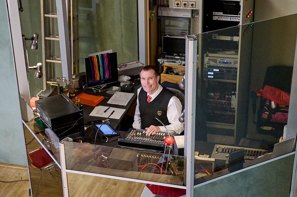 Mann in einem Kontrollraum sitzt an einem Mischpult, umgeben von Technik und Bildschirmen, l&auml;chelnd in die Kamera.