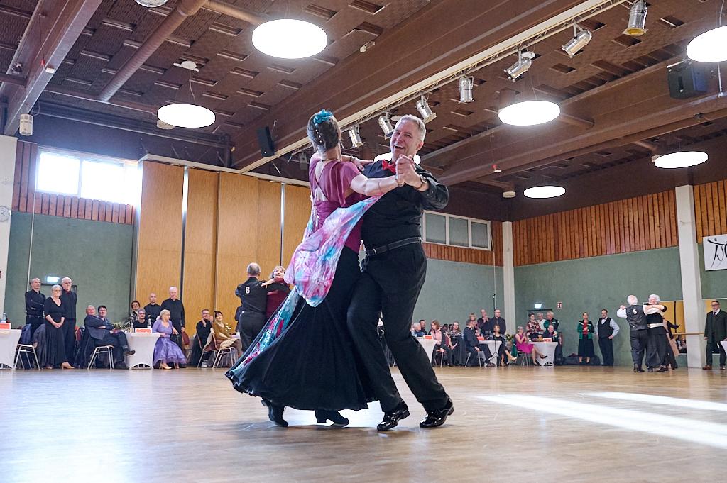 Tanzpaar in eleganter Kleidung tanzt auf Parkettboden vor zuschauendem Publikum in festlicher Atmosph&auml;re.