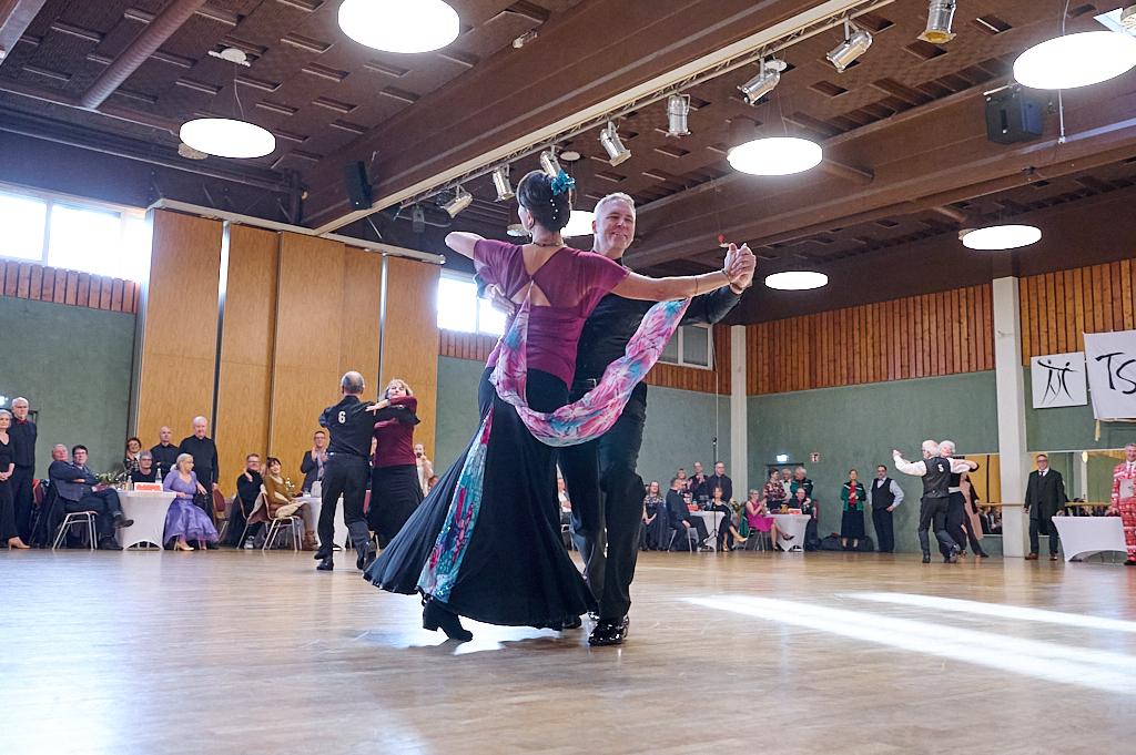 Tanzpaar in eleganter Kleidung tanzt in einem festlichen Saal vor Publikum, umgeben von Tischen und St&uuml;hlen.