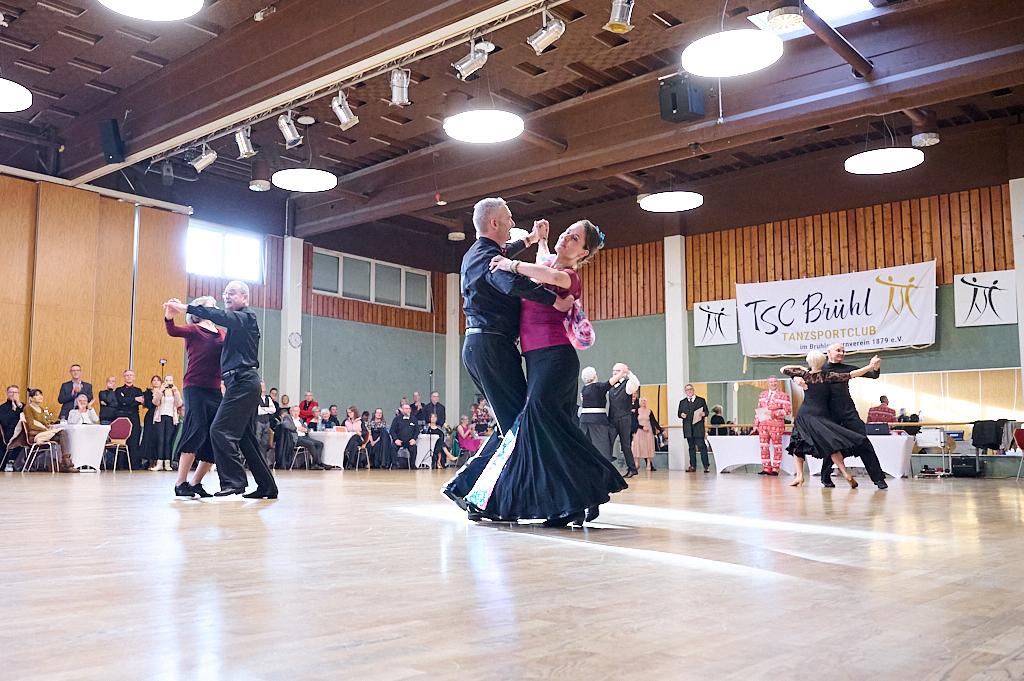 Tanzpaare in eleganter Kleidung tanzen in einem gro&szlig;en Saal mit Tischanordnung im Hintergrund. Banner des TSC Br&uuml;hl sichtbar.