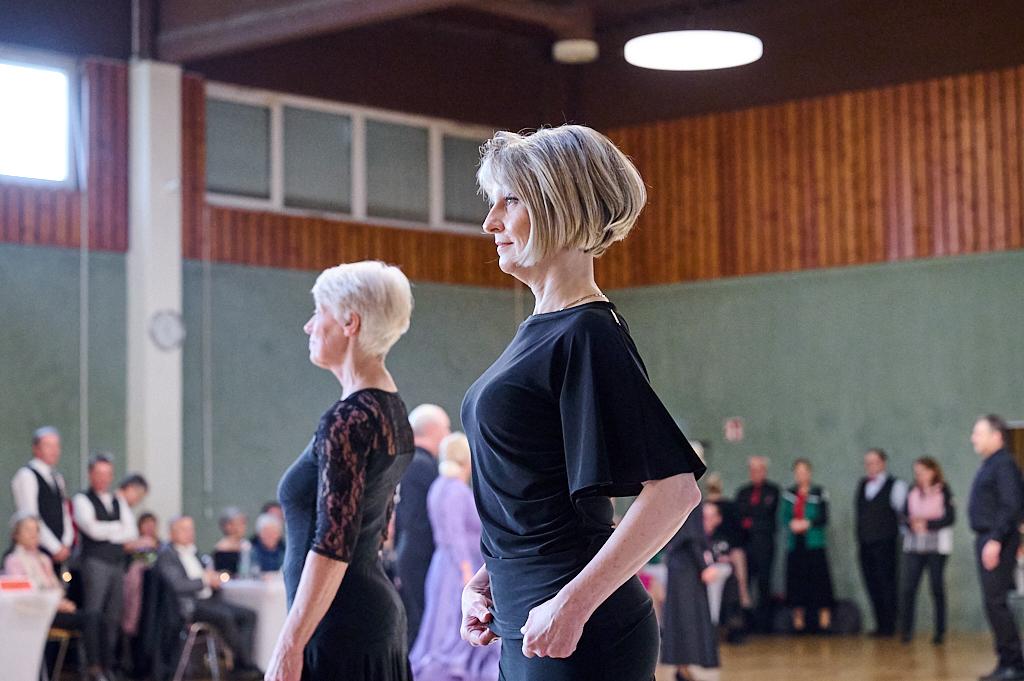 Zwei Frauen stehen in eleganter Kleidung in einer Tanzposition auf einer Turnierb&uuml;hne, Zuschauer im Hintergrund.