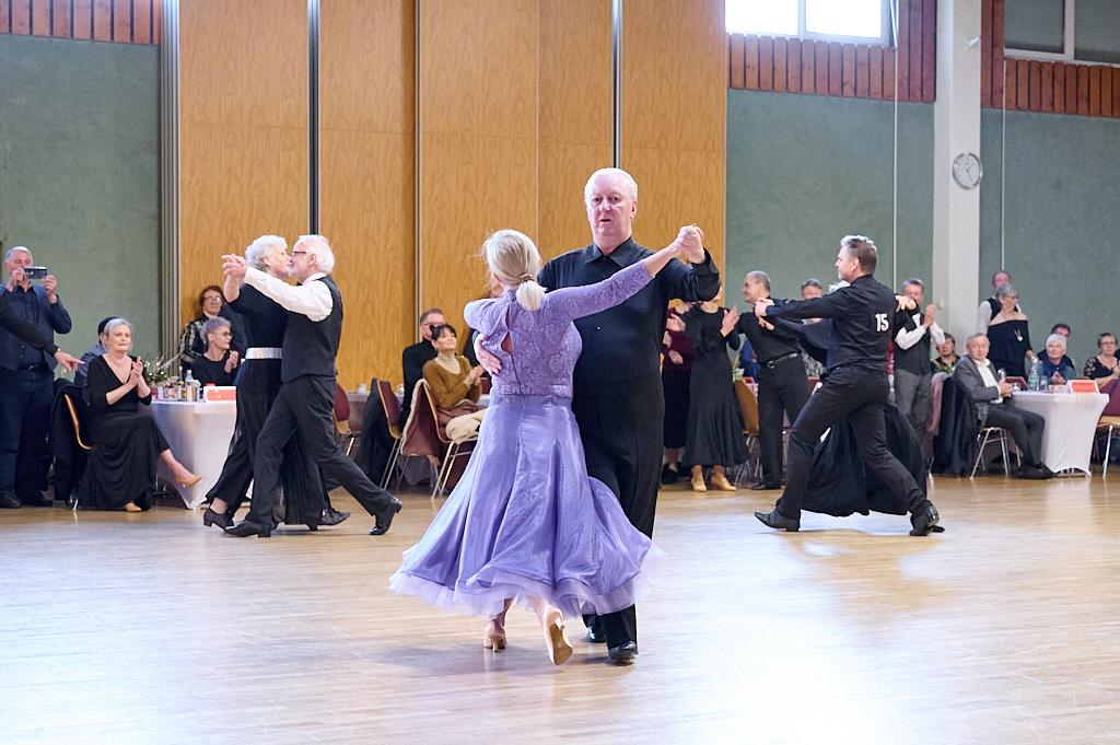Tanzpaar in eleganter Kleidung tanzt in einem festlichen Saal, Zuschauer applaudieren im Hintergrund.