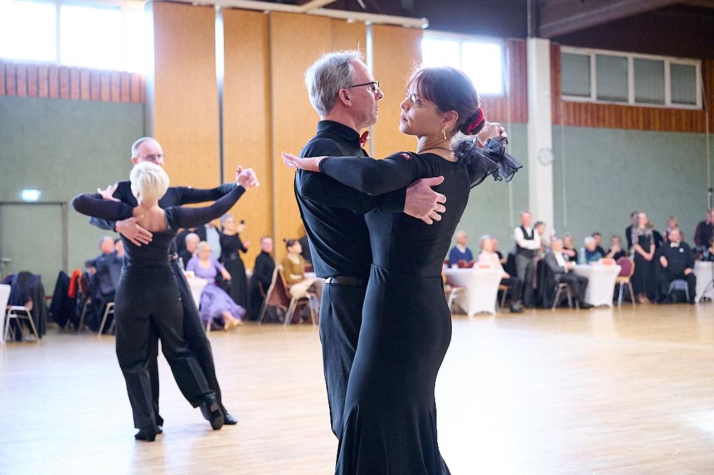 Tanzpaar in eleganter Kleidung bei einer Tanzveranstaltung auf einer Holzb&uuml;hne, umgeben von G&auml;sten.