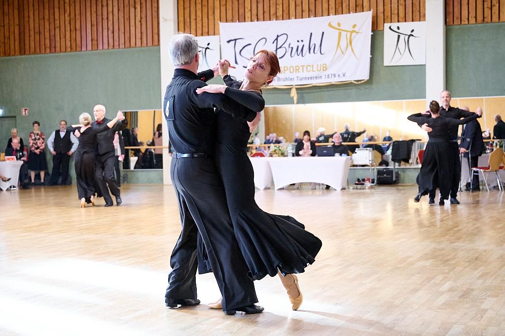 Tanzpaar in eleganter Kleidung tanzt in einem Turnier, Hintergrund mit Zuschauern und Tisch.