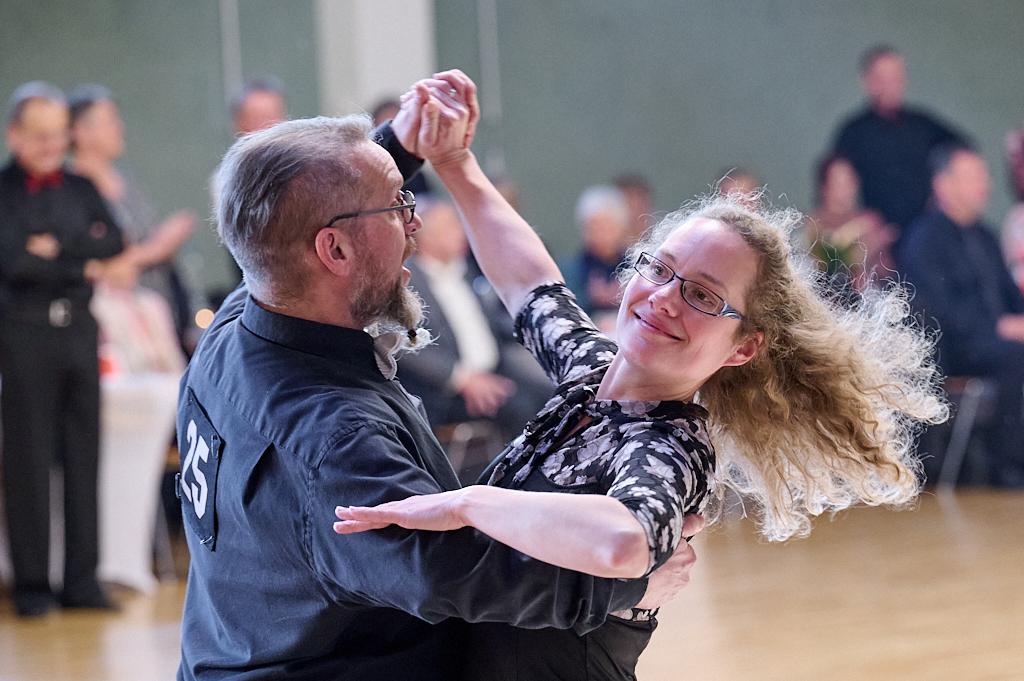 Tanzpaar in eleganter Kleidung tanzt l&auml;chelnd auf einer Tanzfl&auml;che, im Hintergrund Publikum applaudiert.