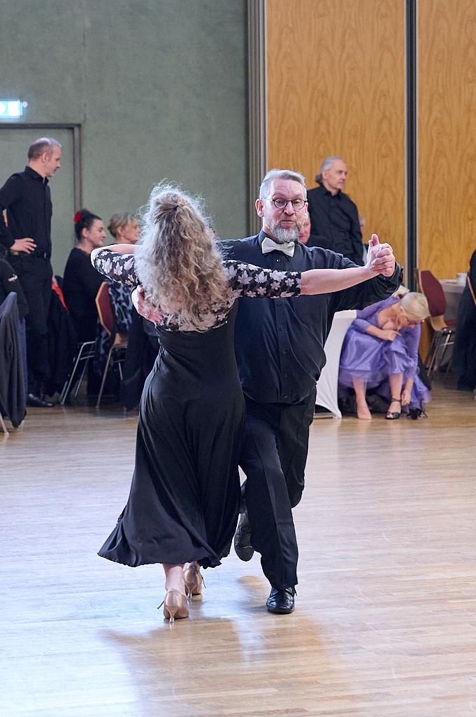 Tanzendes Paar in eleganter Kleidung, umgeben von G&auml;sten in einem festlichen Saal.