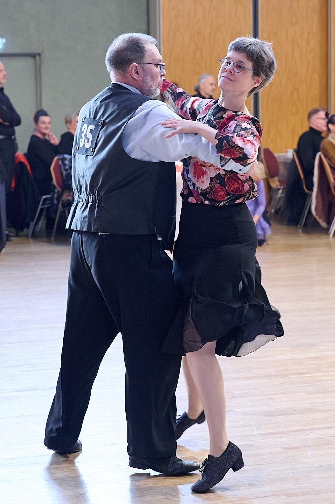 Tanzpaar in eleganter Kleidung beim Tanzen auf einer Tanzfl&auml;che, umgeben von weiteren G&auml;sten im Hintergrund.