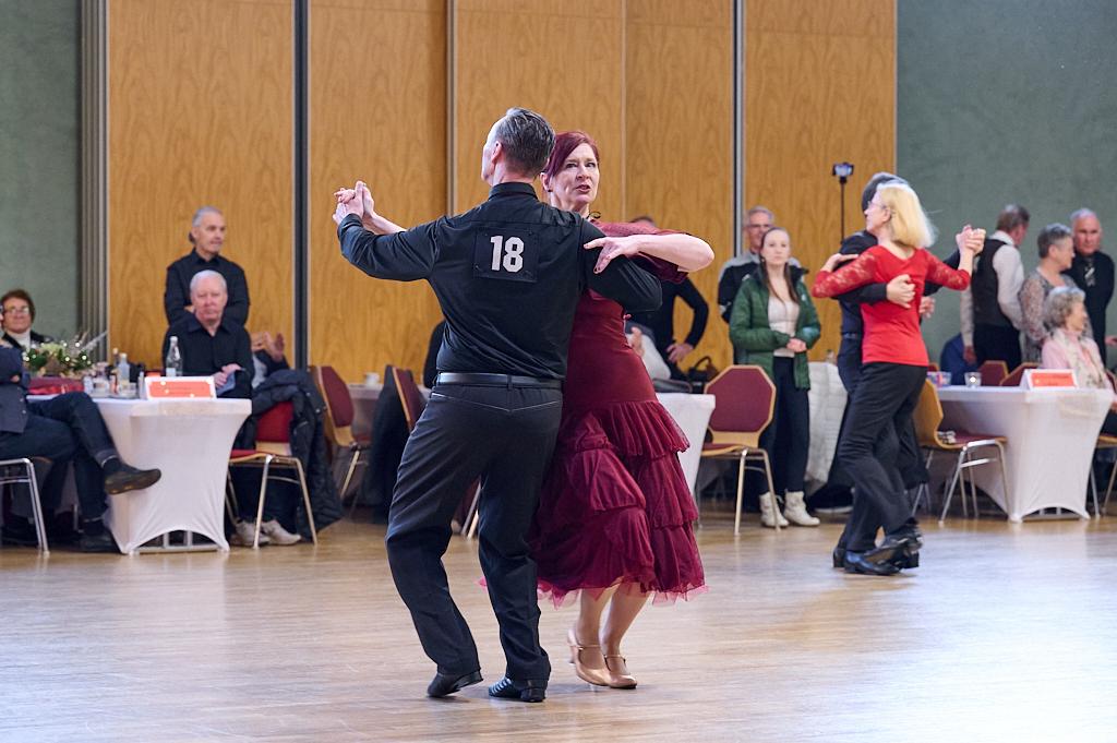 Tanzpaar in eleganter Kleidung, im Mittelpunkt eines Tanzsaals, umgeben von Zuschauern an Tischen.