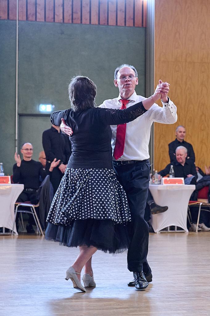 Tanzendes Paar in eleganter Kleidung bei einer Tanzveranstaltung, im Hintergrund applaudierendes Publikum.