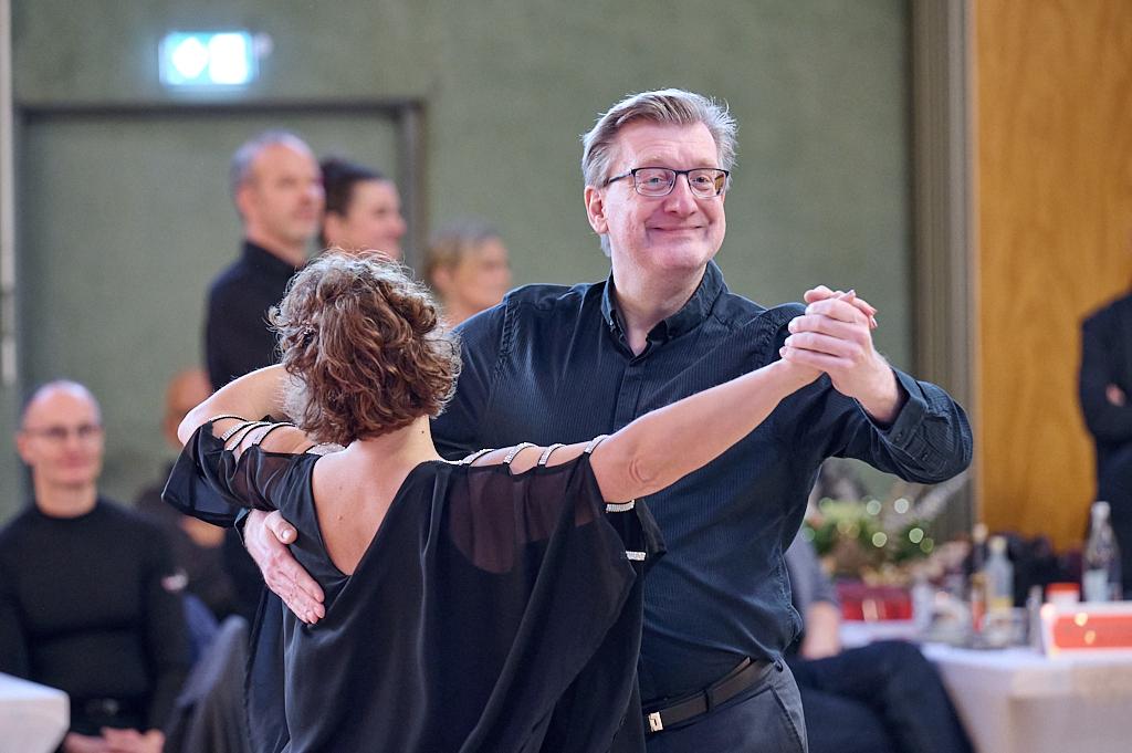 Tanzpaar in formeller Kleidung tanzt l&auml;chelnd in einem festlich dekorierten Saal. Menschen im Hintergrund beobachten das Geschehen.