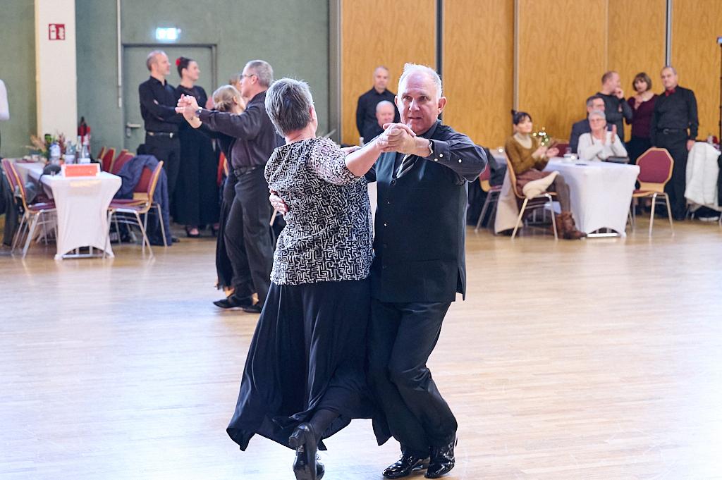Tanzpaar in eleganter Kleidung tanzt auf einer Holzfl&auml;che, w&auml;hrend Zuschauer an Tischen applaudieren.