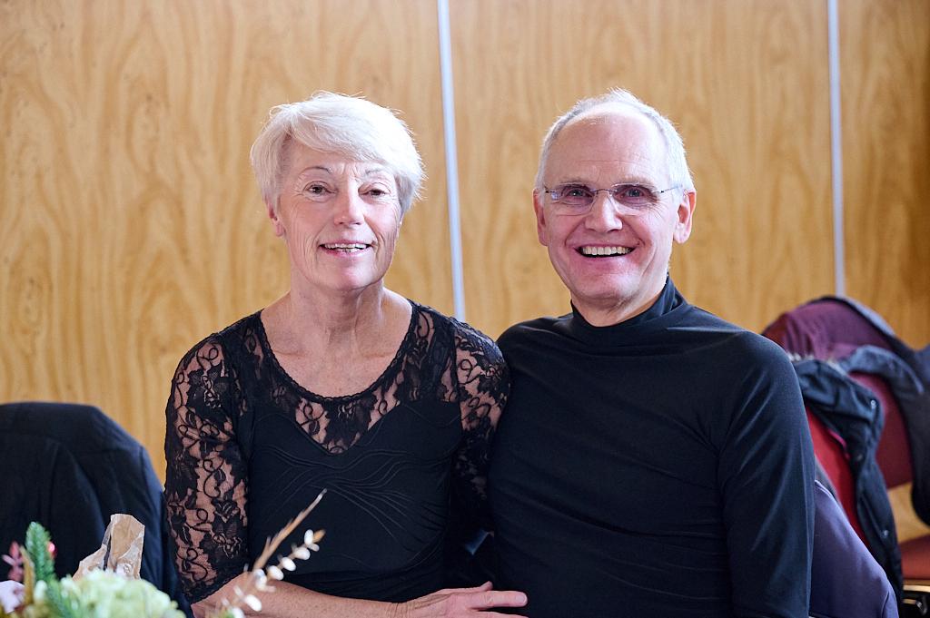 L&auml;chelndes Paar sitzt zusammen an einem Tisch vor h&ouml;lzernem Hintergrund, dekoriert mit Blumen und einem Geschenk.