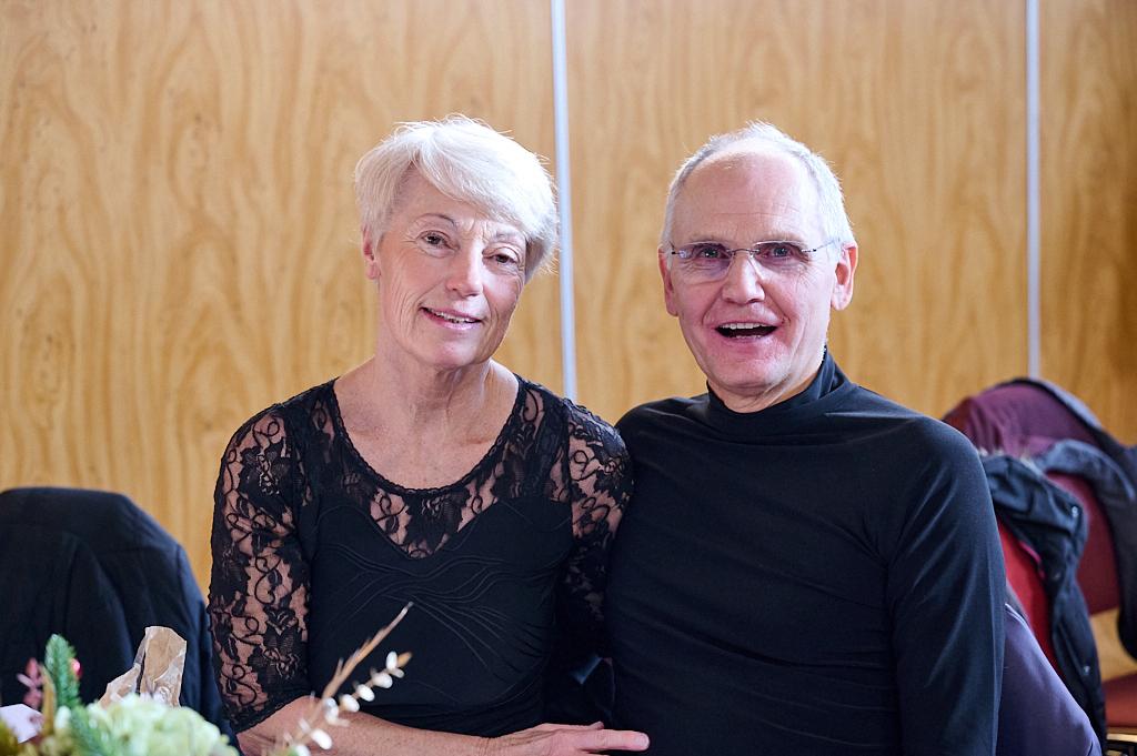 Ein Paar sitzt nebeneinander und l&auml;chelt in die Kamera, vor einem hellen Holzhintergrund.