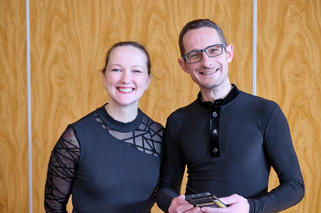 L&auml;chelndes Paar steht vor einer Holzpaneelen-Wand, beide in schwarzen Oberteilen, der Mann h&auml;lt ein Smartphone.