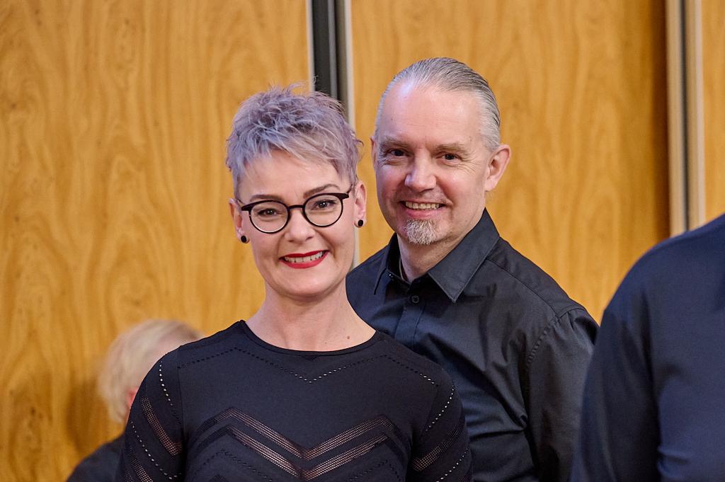 L&auml;chelndes Paar in formeller Kleidung vor einer Holzvert&auml;felung, im Hintergrund sind weitere Personen unscharf zu sehen.