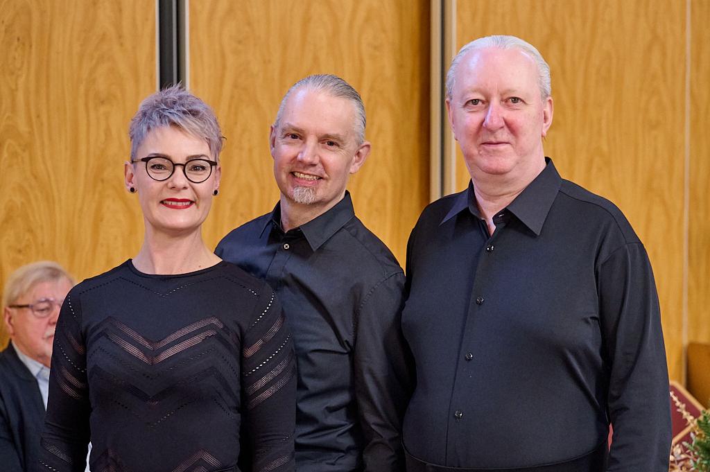 Drei Personen in schwarzer Kleidung stehen l&auml;chelnd vor einer Holzpaneelwand, mit einer Person im Hintergrund.