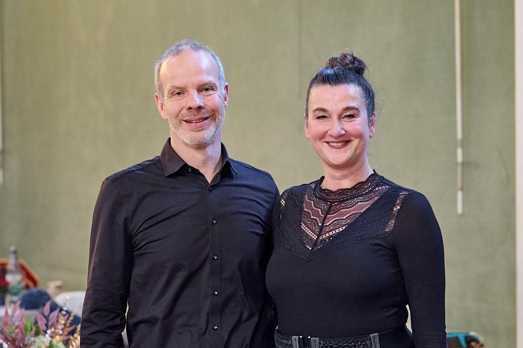 Paar in formeller Kleidung l&auml;chelt in einem Raum mit gr&uuml;nem Hintergrund, im Hintergrund sind Tische und Dekoration sichtbar.