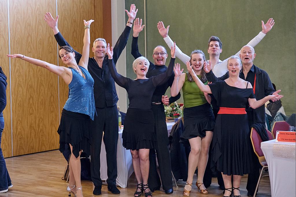 Eine Gruppe von acht tanzenden Personen in eleganter Kleidung hebt die Arme in die Luft und l&auml;chelt fr&ouml;hlich.