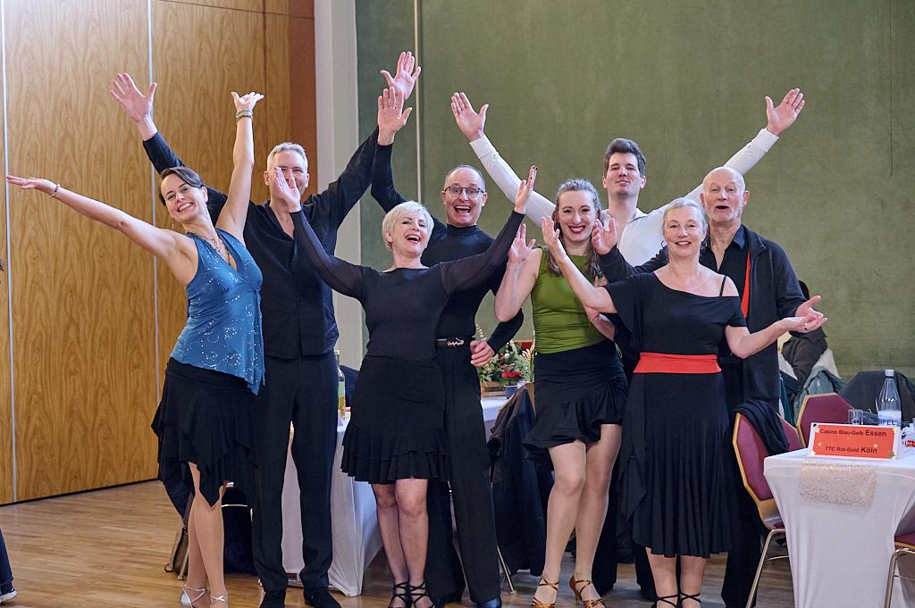 Gruppe von tanzenden Menschen in Schwingkleidung hebt die Arme vor einem festlich gedeckten Tisch.