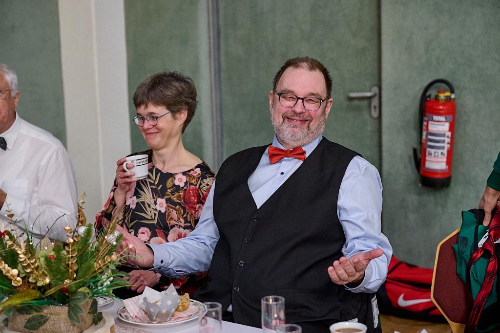 L&auml;chelnder Mann mit Brille und Fliege sitzt an einem Tisch, w&auml;hrend eine Frau mit Kaffeetasse neben ihm steht.