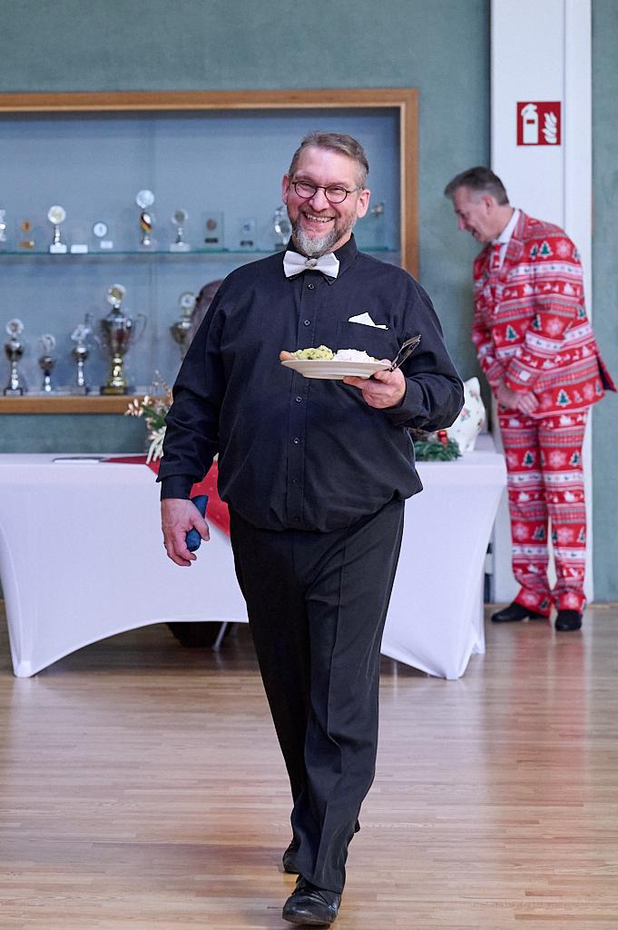 L&auml;chelnder Mann in schwarzem Hemd und Fliege tr&auml;gt einen Teller mit Essen, w&auml;hrend ein anderer Mann im Hintergrund steht.