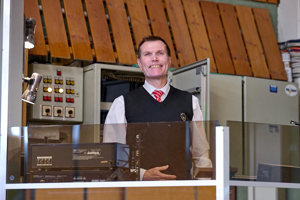 L&auml;chelnder Mann in Weste hinter einem Empfangstresen mit Technik und Steuerungselementen im Hintergrund.