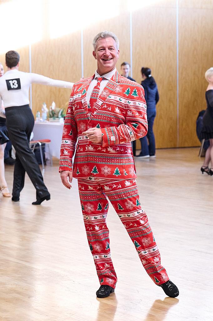 Mann in einem weihnachtlich gemusterten Anzug l&auml;chelt auf einem Tanzparkett, im Hintergrund T&auml;nzer und festliche Dekoration.
