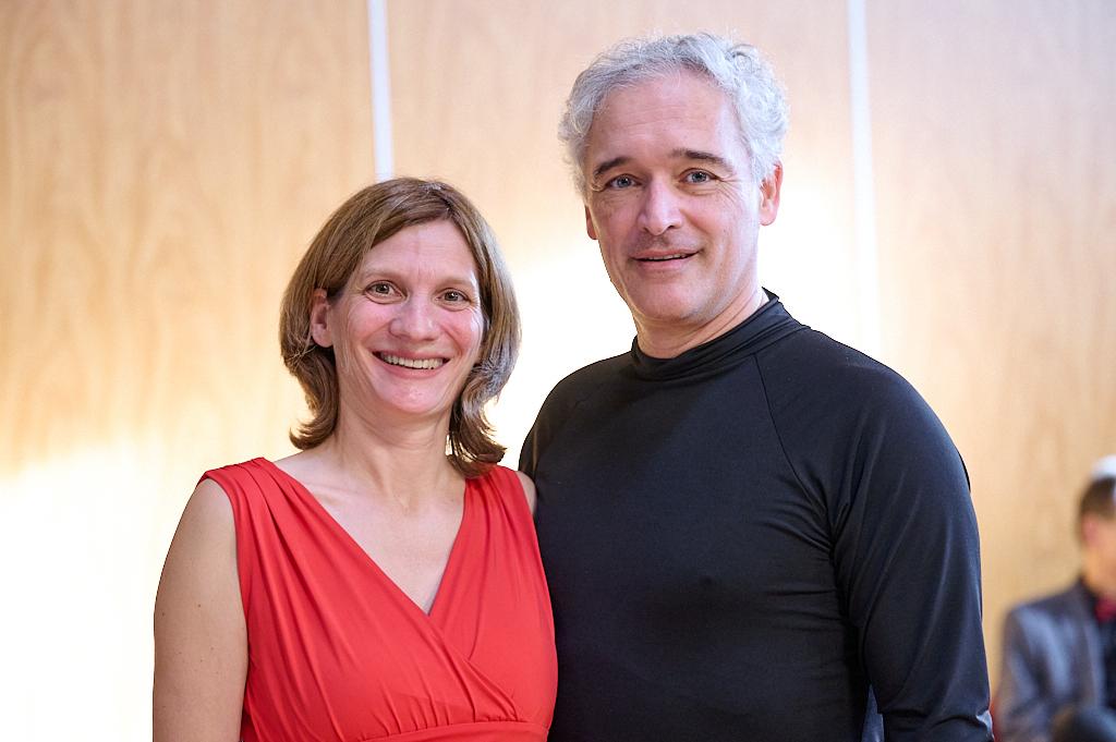 Portrait eines l&auml;chelnden Paares, sie in rot, er in schwarz, vor einer hellen Holzoberfl&auml;che.