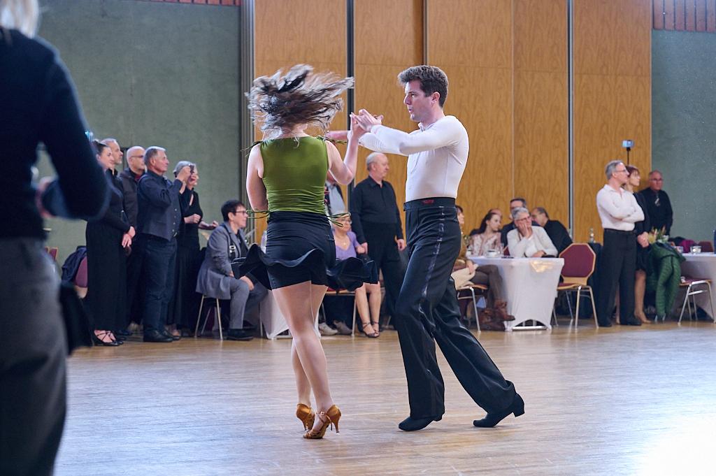 Tanzpaar in Bewegung auf der Tanzfl&auml;che, Zuschauer im Hintergrund, festliche Atmosph&auml;re in einem Saal.
