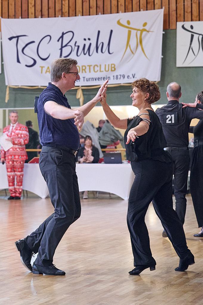 Tanzpaar in formeller Kleidung tanzt fr&ouml;hlich in einem Sportclub, im Hintergrund Zuschauer und ein Banner des Clubs.